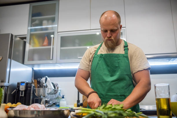 a blond man with a beard and a green apron prepares dinner in his home kitchen. - routine foods and drinks clothing household equipment imagens e fotografias de stock