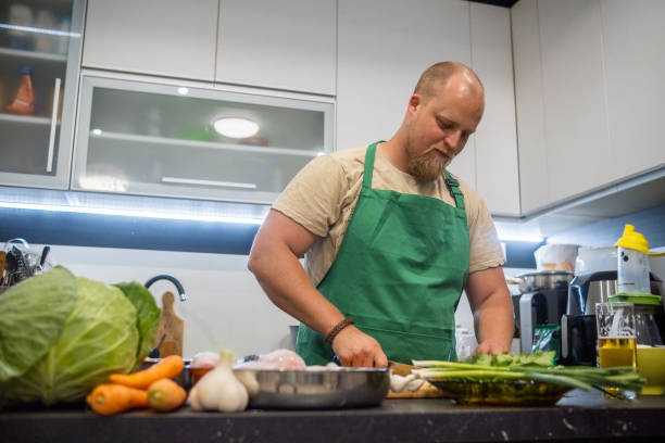 a blond man with a beard and a green apron prepares dinner in his home kitchen. - routine foods and drinks clothing household equipment imagens e fotografias de stock