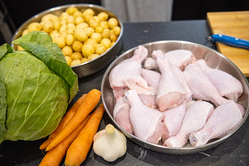 Dinnerware on the kitchen counter. A tray full of chicken legs. Onions, carrots, cabbage, and a pan filled with baby potatoes.