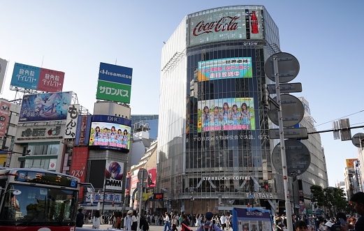May 24, 2023 - Shibuya City, Japan: Buses and pedestrians make their way along Dogenzaka outside Shibuya Station. Commercial signs and projection screens line the walls by Shibuya Crossing with a  colorful display in the late afternoon.