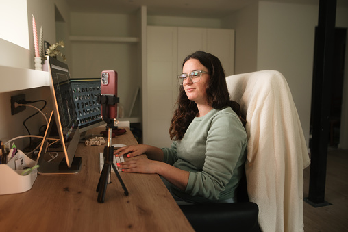 A young woman in a desk with a computer, looking at a smart phone on a tripod - video call, streaming concept