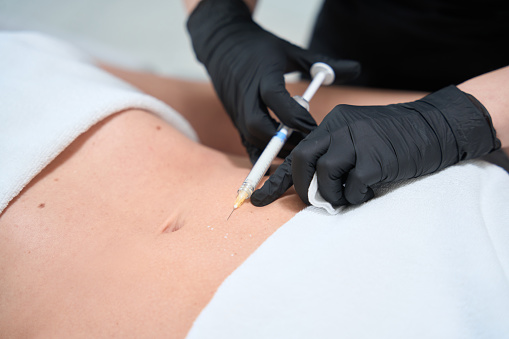 Employee of a cosmetology clinic makes injections in the stomach of a client, an esthetician works in protective gloves