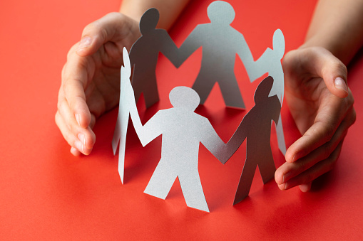 Paper people surrounded by hands in gesture of protection on red background.