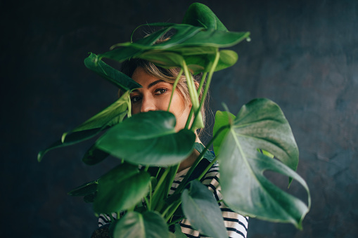 A portrait of a Caucasian female hiding behind a plant while standing against grey background.