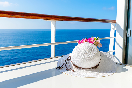 A woman's white sun hat sits next to a small bouquet of flowers on a cruise ship balcony with the blue sea in view on a summer day.