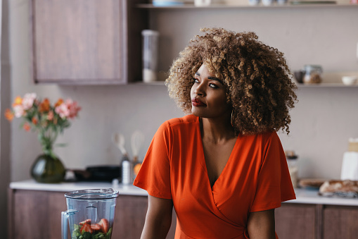 A portrait of a young black woman in an orange dress standing in a bright kitchen.