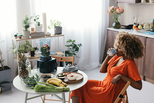 A happy black woman in an orange dress sitting at a round kitchen table, enjoying a cup of coffee.
