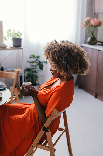 A happy black woman in an orange dress sitting at a kitchen table, enjoying a cup of coffee.