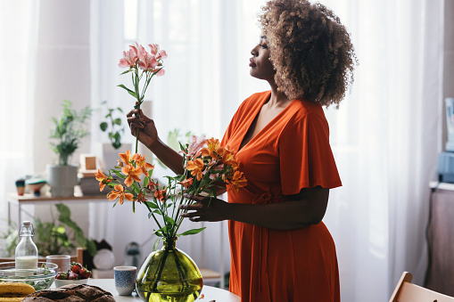 A happy black woman in an orange dress arranging a bouquet of flowers on the kitchen table.