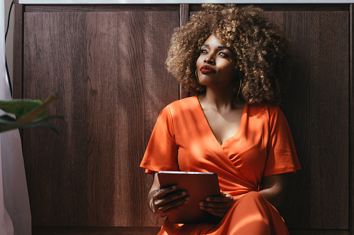 A portrait of a happy African American woman in an orange dress sitting on the floor, using a tablet.