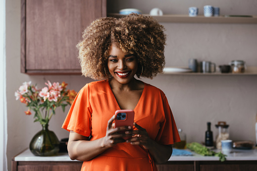 A portrait of a happy black woman standing in the kitchen, using her mobile phone.