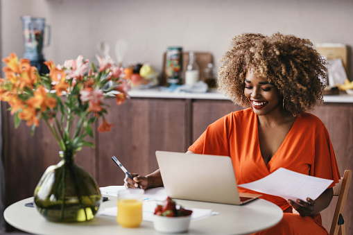 A beautiful black female entrepreneur working from home, sitting at the kitchen table and analyzing a report on her computer.