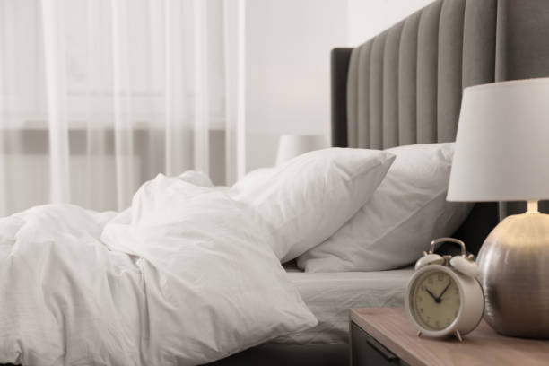 istock Lamp, alarm clock on bedside table and comfortable bed in light room. Stylish interior 1500091318