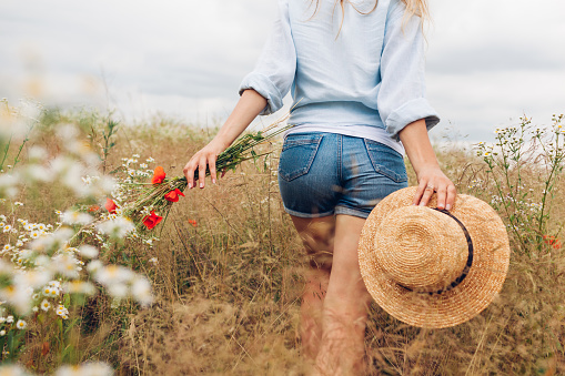 Rustic summer female accessory and clothes. Woman holding straw hat wearing linen shirt and shirts walking in meadow picking wildflowers.