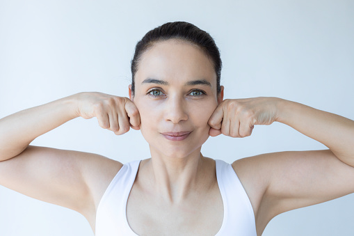 Caucasian female is doing face yoga gymnastics for non-surgical rejuvenation and is looking at camera in front of white background. Anti aging concept.