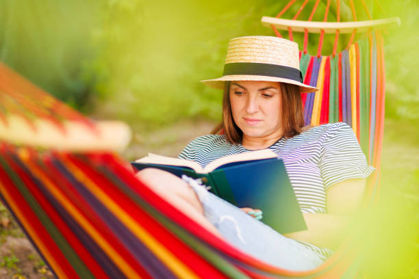 istock Young woman reading book while lying in comfortable hammock at green garden 1500099876