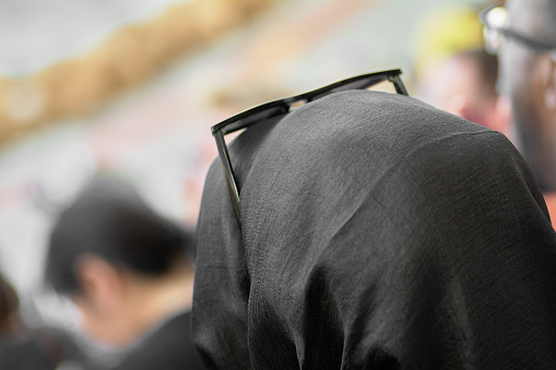 Unrecognizable woman with black headscarf and black glasses in the foreground and people out of focus in front of her.