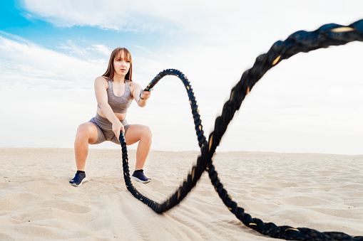Muscular young concentrated woman working out with battle ropes on the beach, intense functional circuit training. Doing sport outdoors. gym, fitness and workout concept. Selective focus