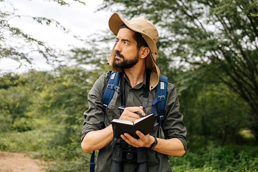 Man in nature with hat, backpack, and binoculars taking field notes