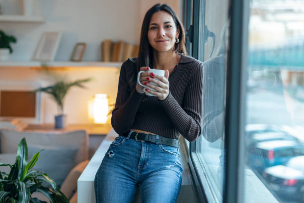 istock Beautiful thoughtful woman drinking a cup of coffee while looking at camera next to the window at home 1500107971