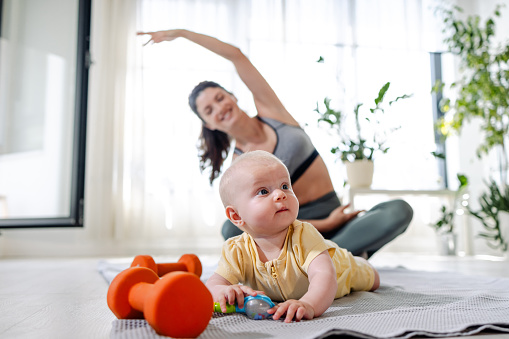 Portrait of a cute baby playing on the floor while her mom is sitting in the lotus position and stretching