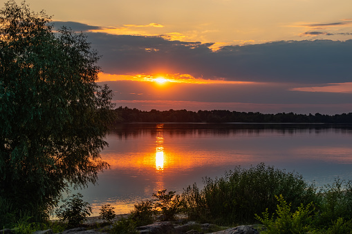 A picturesque sunset with clouds over the river, trees and grass on the shore, the reflection of the Sun on the water. Nature on a summer evening