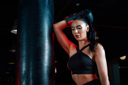Sportswoman in boxing gloves leaning on punching bag in gym.
