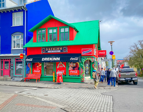 Reykjavik, Iceland-May 25, 2023 -Locals and tourists walk on the sidewalk past the colorful red and green Drekinn fast food restaurant with painted dragons and Coca-Cola ads in the windows.