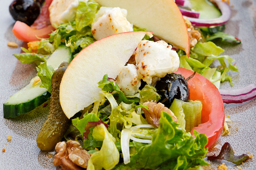 Close-up of a fresh salad with grilled goat cheese, apple, celery, walnuts, tomato, red onion, lettuce, cucumber, pine nuts, walnuts, olives and pickle topped with a honey dressing on a gray plate.
