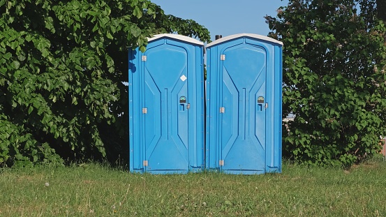 Pair of Blue Plastic Mobile Public Toilet Outhouse Standing in the Middle of High Grass Meadow During Open Air Event Music Festival