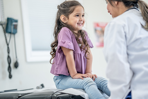 A sweet little girl sits up on an exam table during a doctors appointment.  She is dressed casually and her female doctor is seated in front of her as they talk.