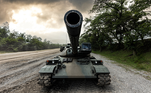 A wide-angle front view of a Japanese self-defense force military tank driving down a dirt road lined with trees with a dramatic sky.