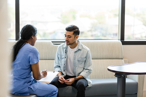 The young adult male sits on the hospital room couch to talk with the hospital insurance specialist to update medical information.