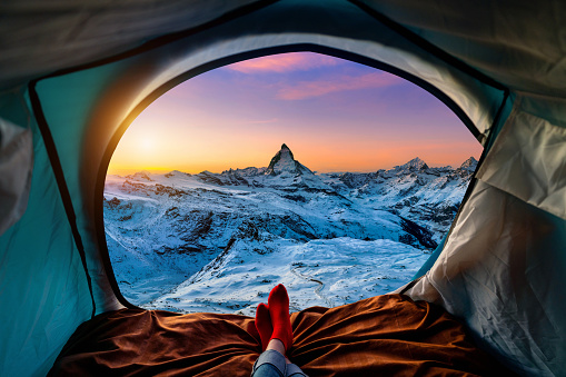 Woman cross leg on blanket in camping tent with sleeping bags on mountain hill. view from inside with Matterhorn mountains.