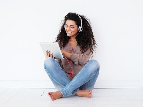 african american young woman sitting on floor with tablet pc computer and headphones listening to music at home
