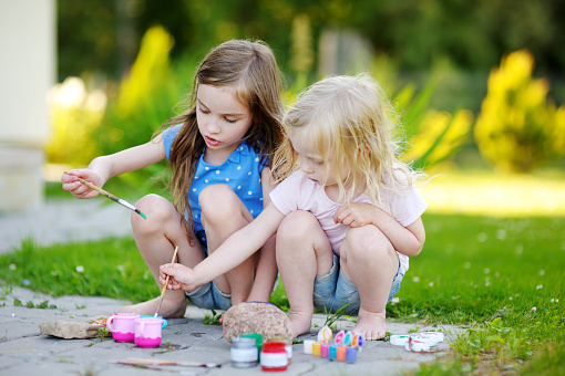 Two cute little sisters painting on stones together on hot summer day