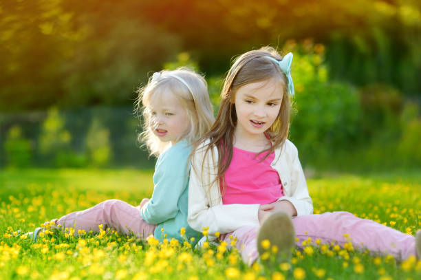 Rear view of two little girls, both with brown hair, sitting in a garden, embraced, having bonding moments. They are looking over shoulder, smiling at camera.
