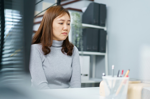 Portrait of unhappy - sad Asian female employee sitting in the office with her negative expressing. Stressful Asian woman feels uncomfortable during working in the negative working environment.