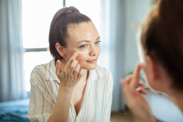 young woman putting on makeup while looking in a mirror at home - mirror women bathroom make up imagens e fotografias de stock