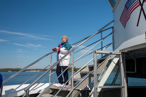 Senior Chinese tourist holding a camera in hand on cruise ship on Saint Lawrence river. America flag printed on the ship. Thousand Islands. Canada.