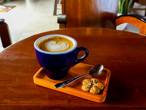 Enjoying hot cappuccino with heart shaped latte art in the morning at traditional living room, served in a blue cup with cookie on a wooden tray and placed on round wooden table