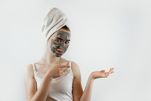 beautiful girl in towel applying black caly mask on face, anti aging beauty treatment isolated on white gray. Skin care