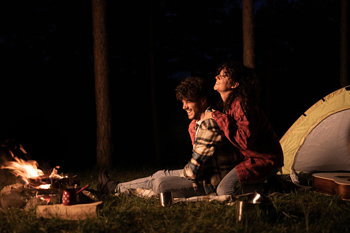 Young happy couple relaxing by the fire during camping night in nature. Copy space.