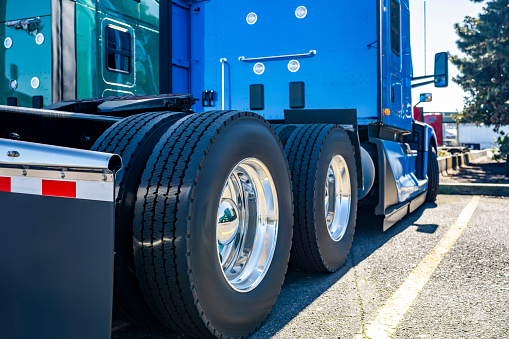 Industrial standard blue big rig semi truck tractor with new wheels with endurance tires on polished durable alloy rims standing on the truck stop parking lot beside with another semi trucks