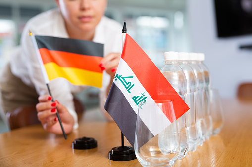 Unrecognizable woman preparing room for international negotiations and communication discussions of leaders. Lady sets miniatures flags of Iraq and Germany on table. Unfocused shot