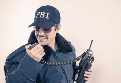 White FBI agent man talking on radio, calling in for backup during a crises, wearign a heavy blue coat, baseball cap, vintage mustache, and colored shades.