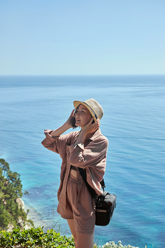 woman posing in front of ocean with hat, in the style of earth tone color palette, mediterranean landscapes, mountainous vistas, pink and azure, utilitarian, precisionist, chic