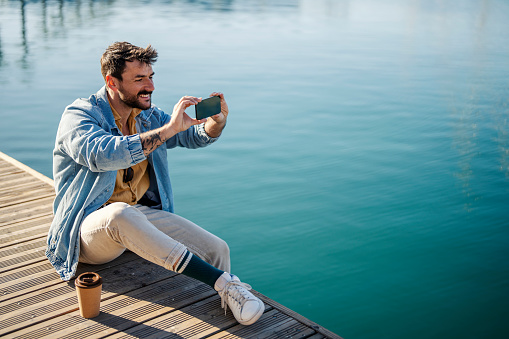 A cheerful man is capturing self portraits while sitting on the pier.