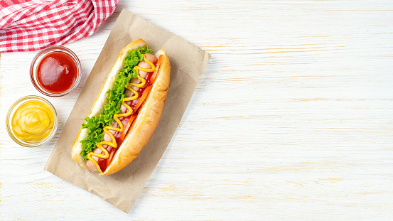 Homemade Hot Dog with yellow mustard, ketchup, tomato and fresh salad leaves on white wooden background. Fast food, street food, american cuisine. Top view, banner, header with copy space