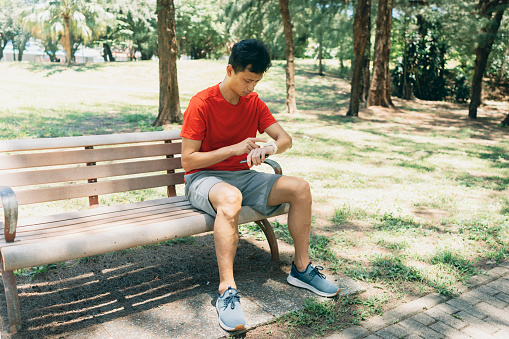 An Asian man in a natural environment on a summer day, getting ready to start his jog. He checks his watch, preparing to synchronize it with a mobile app to record his exercise progress and data.Wearable technology. Healthy living lifestyle, sports routine concept